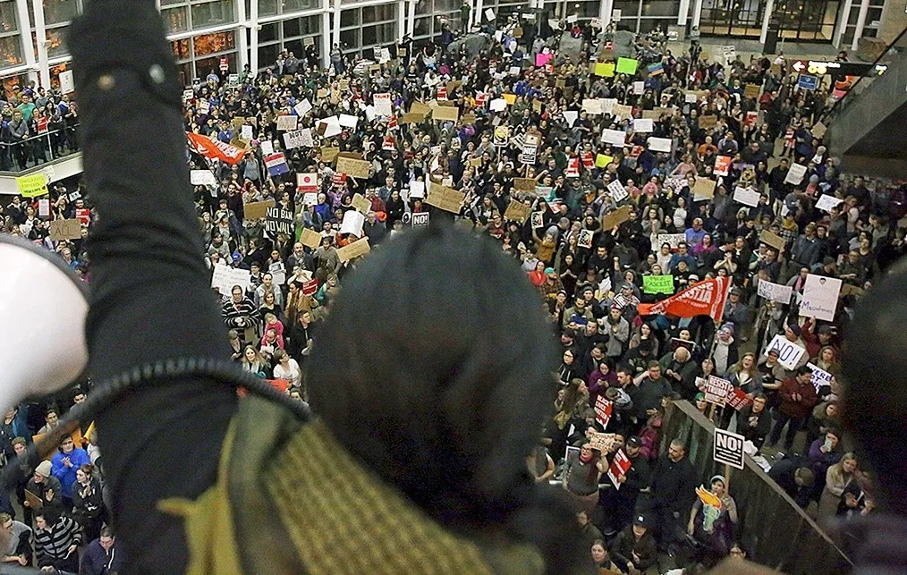 A speaker at a rally, back to camera, raises their fist to hundreds of protestors in an airport